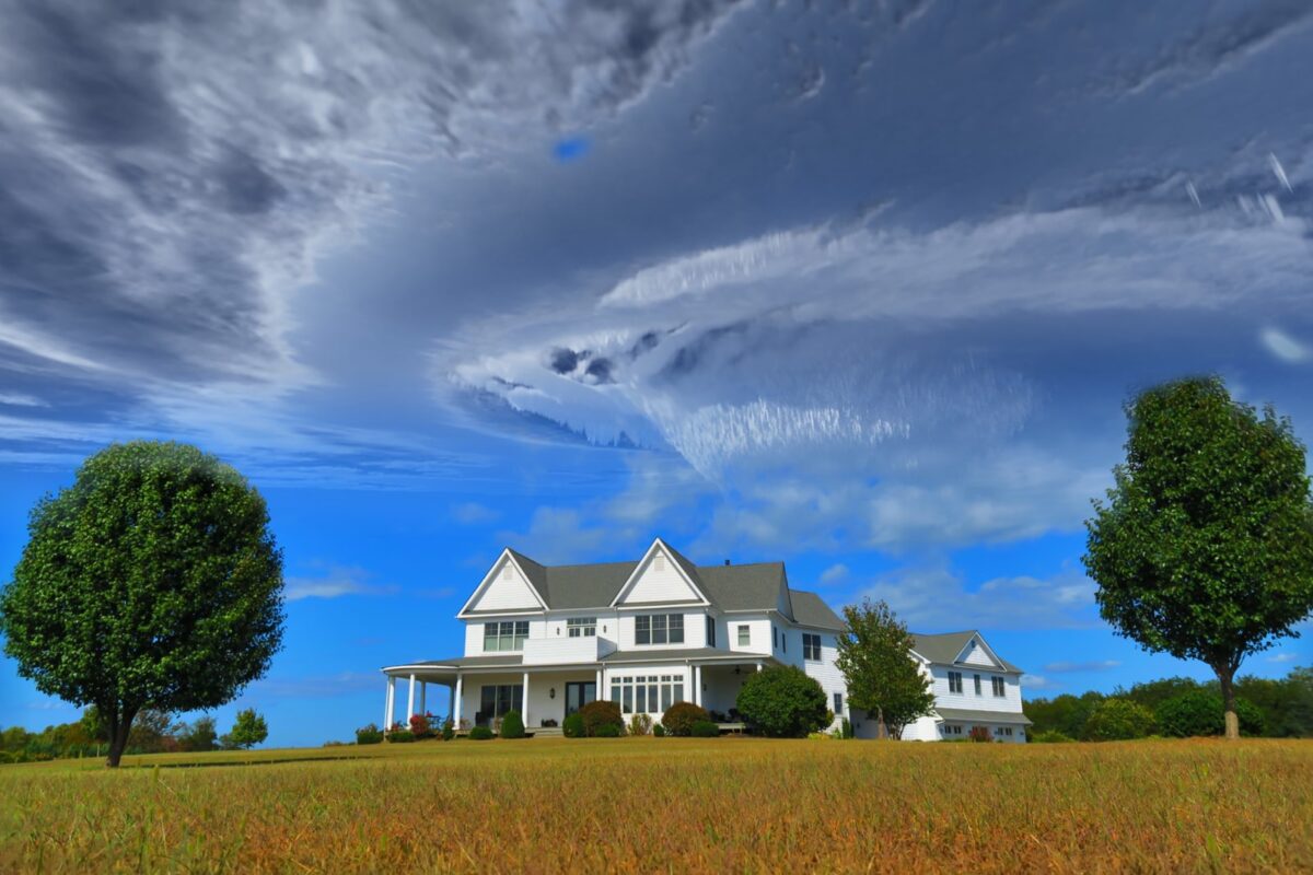 white and gray house between two green tall trees under gray clouds forming swirl during daytime